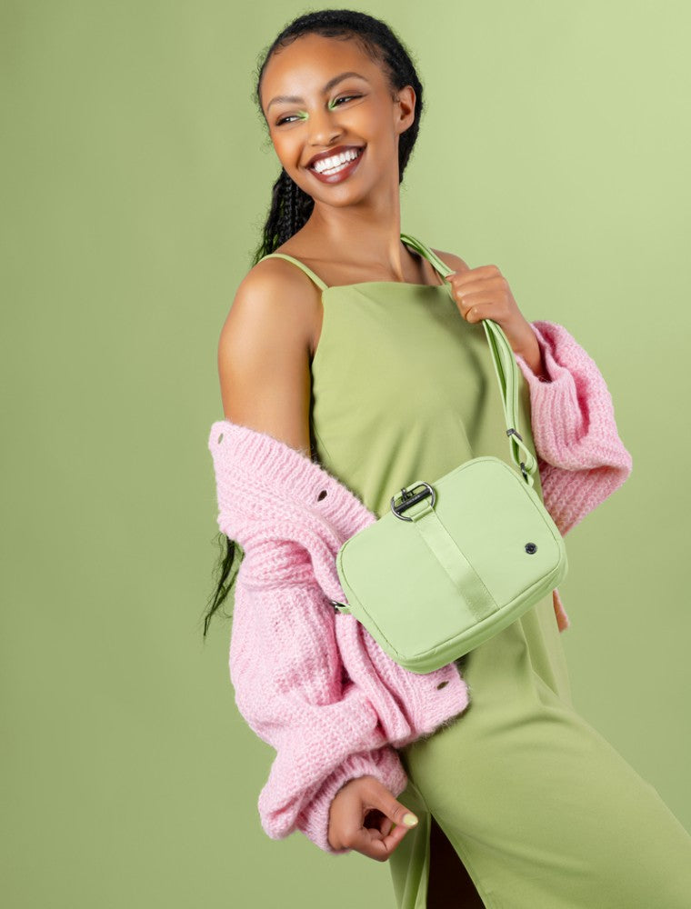 Woman wearing a green dress and pink knitted cardigan, smiling while holding a light green square-shaped Pacsafe anti-theft crossbody bag made with ECONYL® regenerated nylon, featuring a clasp and attachable strap.