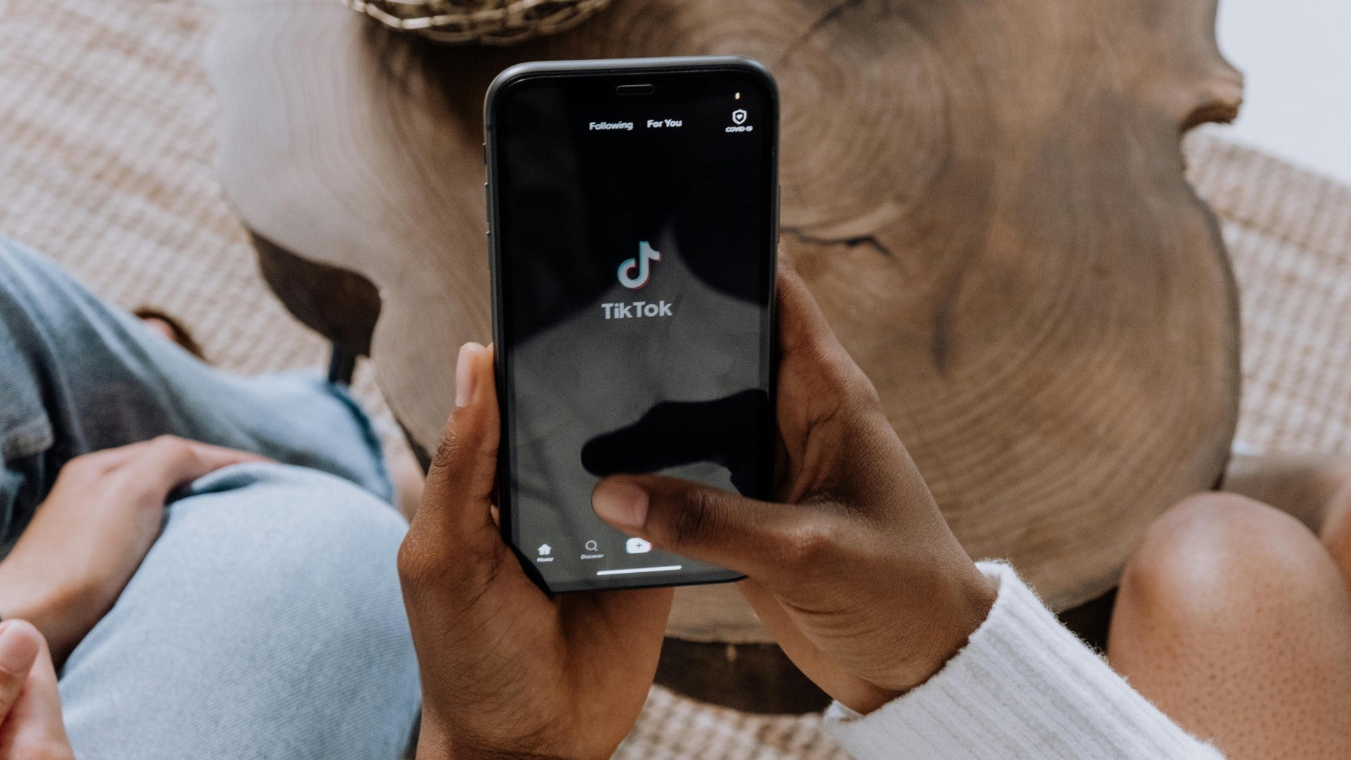 Person holding a smartphone displaying the TikTok app loading screen, seated near a wooden coffee table.