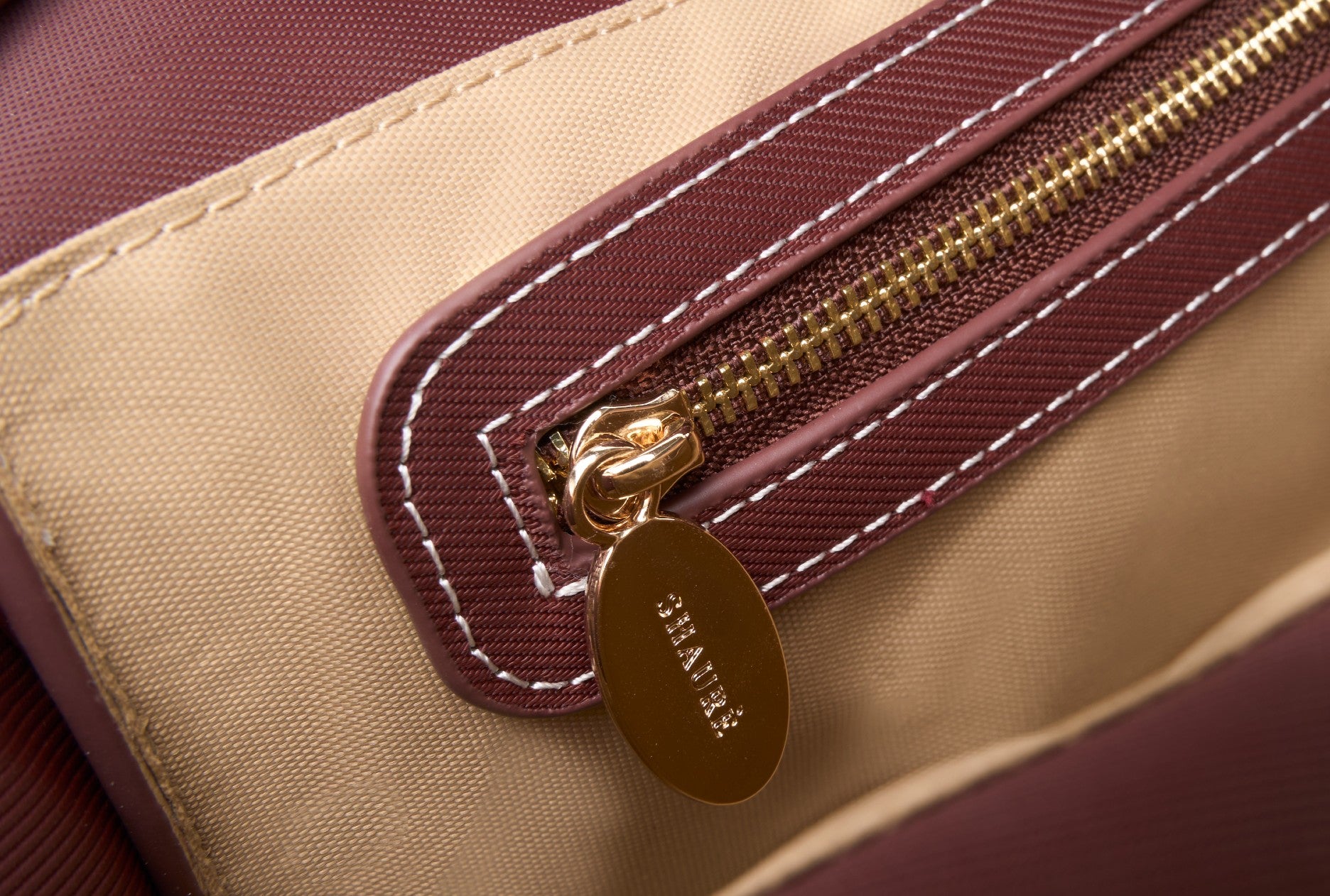 Close-up of a beige and burgundy bag, featuring ECONYL® regenerated nylon, with a gold zipper and 'Chantecler' tag. 