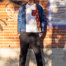 Load image into Gallery viewer, Man standing against a brick wall with graffiti, wearing a blue jacket made with econyl nylon, white shirt, and patterned scarf.
